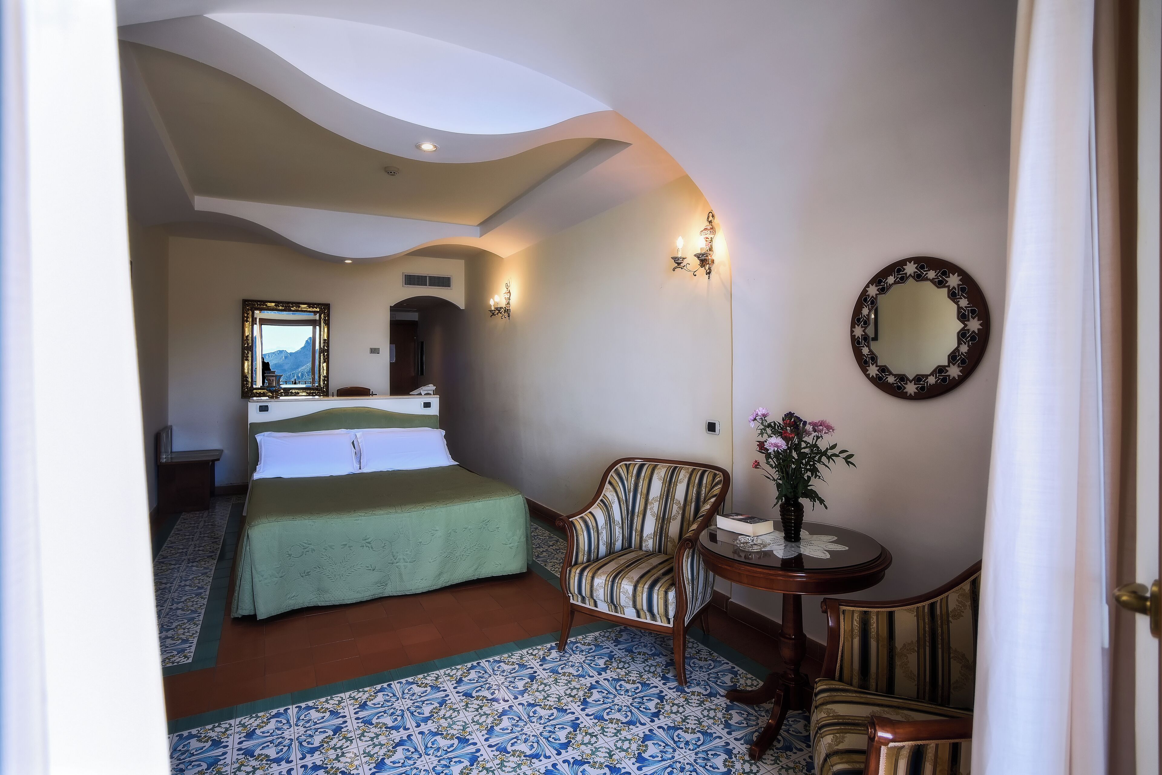A hotel room with a bed, a chair, a small table with a vase, and a mirror on the wall.