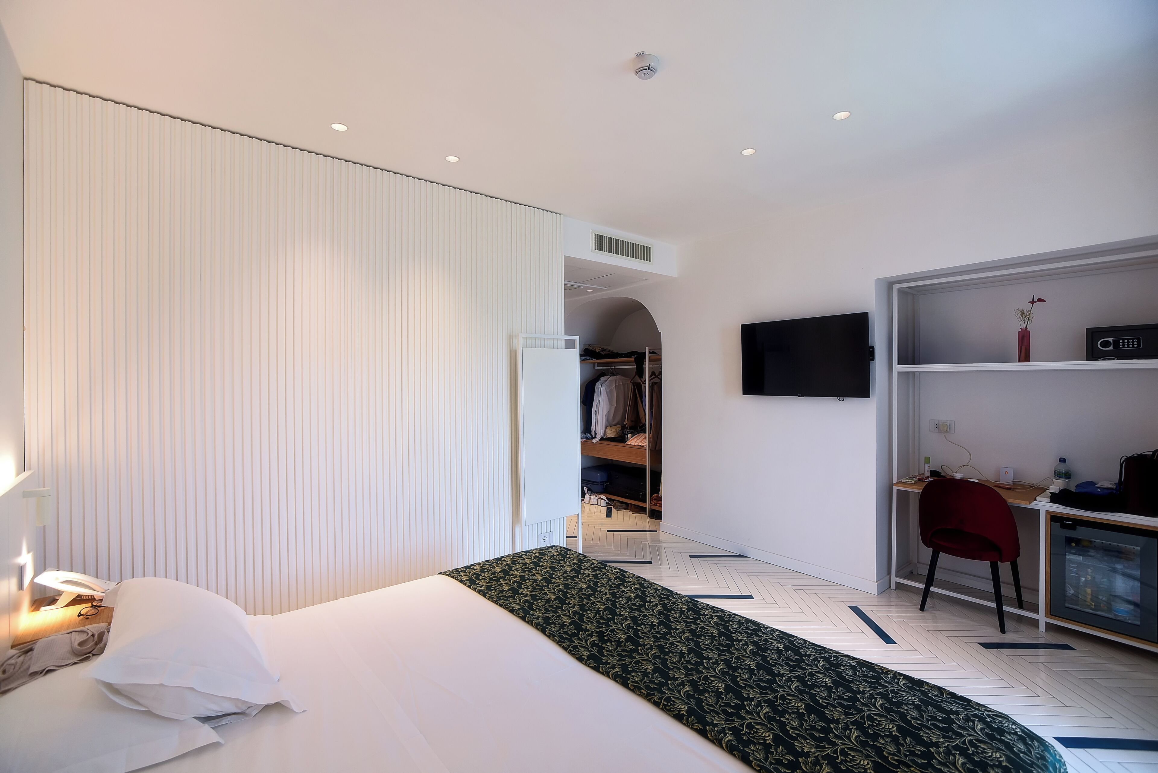 A hotel room with a bed, bedside table, lamp, wardrobe, desk with chair, and a TV mounted on the wall.