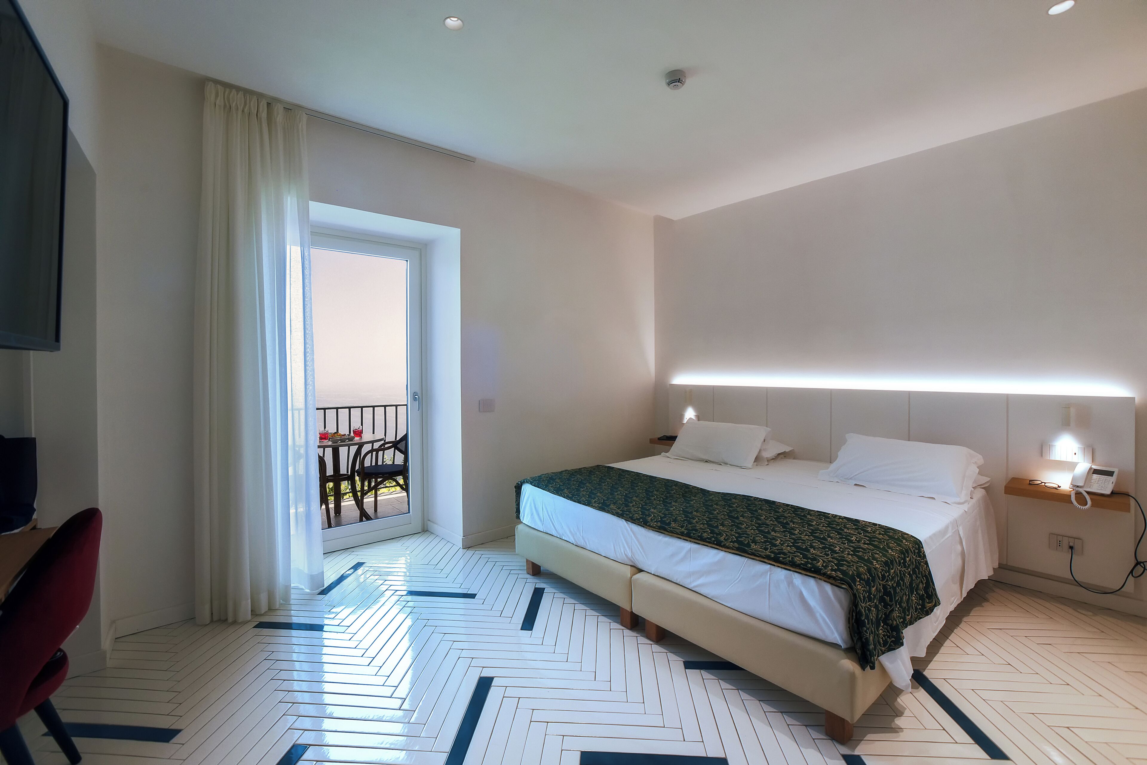 A hotel room with a bed, bedside table, lamp, and a view of the balcony.