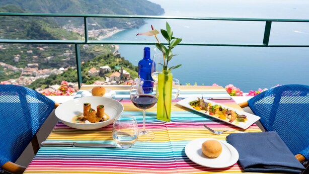 A table set with plates of food, a wine glass, and a vase with a plant, overlooking a coastal view. - Hotel Graal (Ravello)