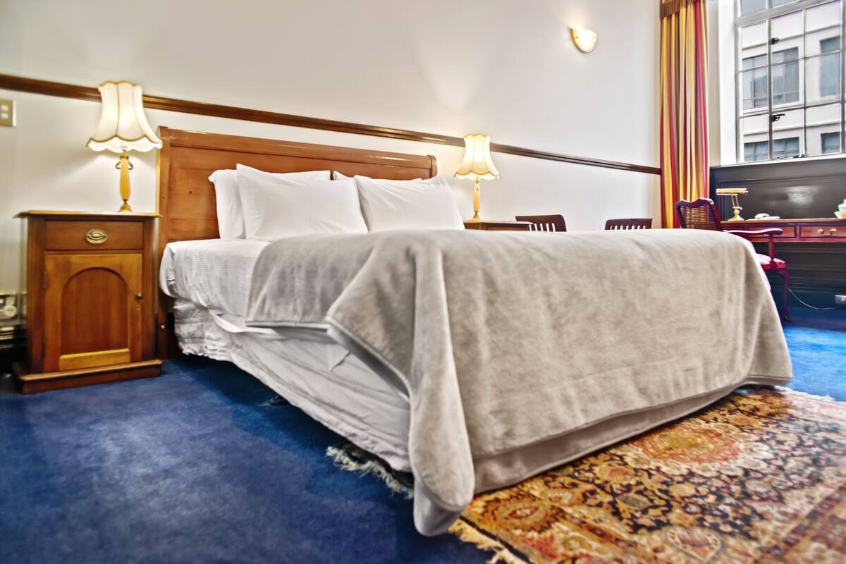 A neatly made bed with a wooden headboard, two bedside tables with lamps, a patterned rug, and a window with curtains.