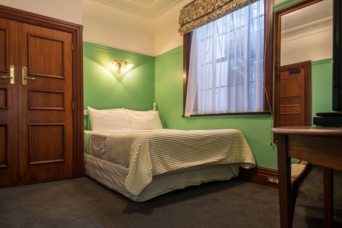 A bedroom with a bed, a nightstand, a mirror, and a window with curtains.