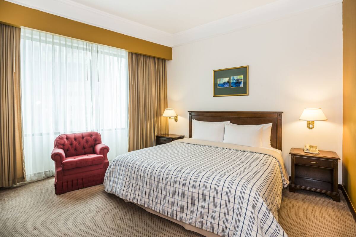 A hotel room with a large bed, a red armchair, two bedside tables with lamps, and a painting on the wall.
