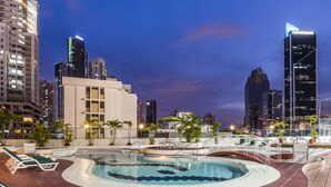 Una piscina en la azotea con sillones de descanso y vistas al horizonte de la ciudad al anochecer.