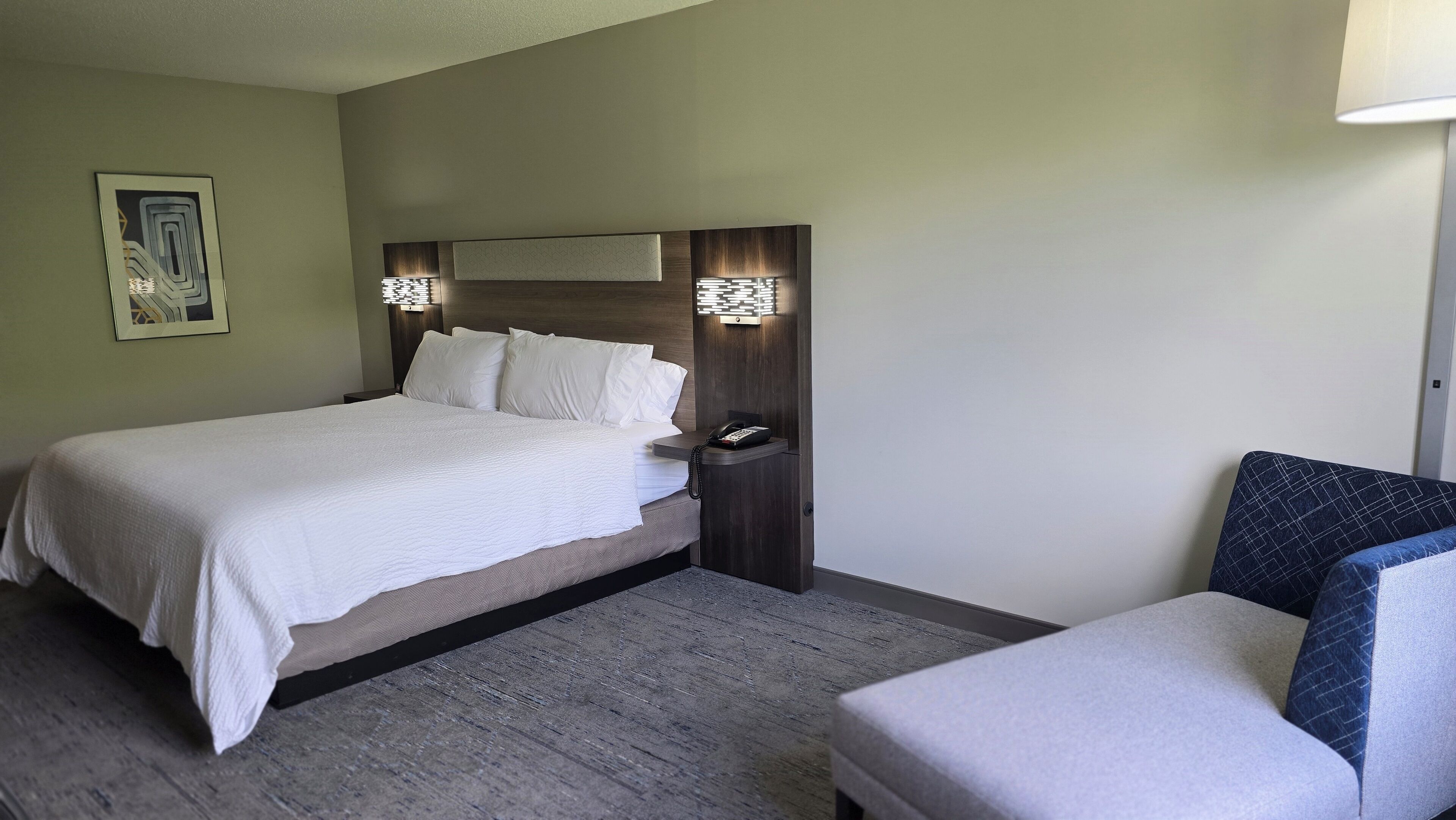 Quarto de hotel com cama, mesa de cabeceira, luminária e uma cadeira.