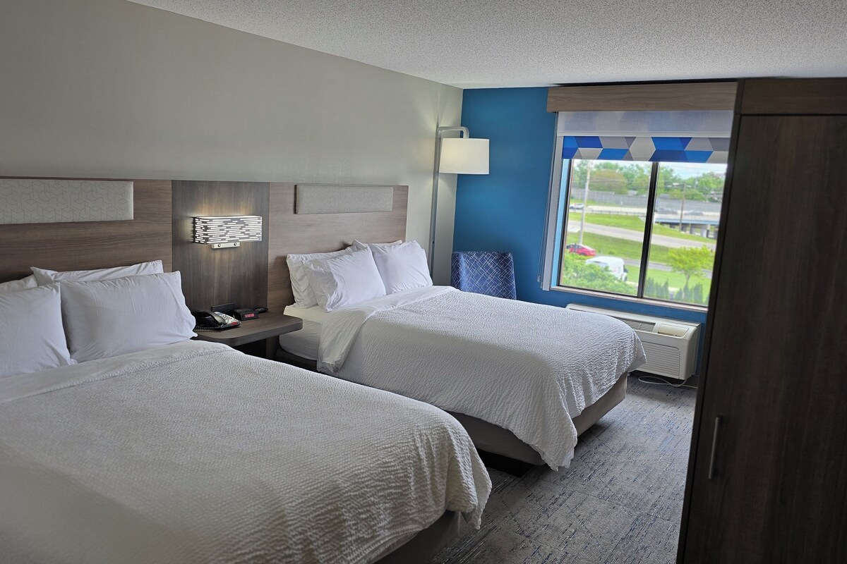 A hotel room with two beds, a nightstand, a lamp, and a window with a view.