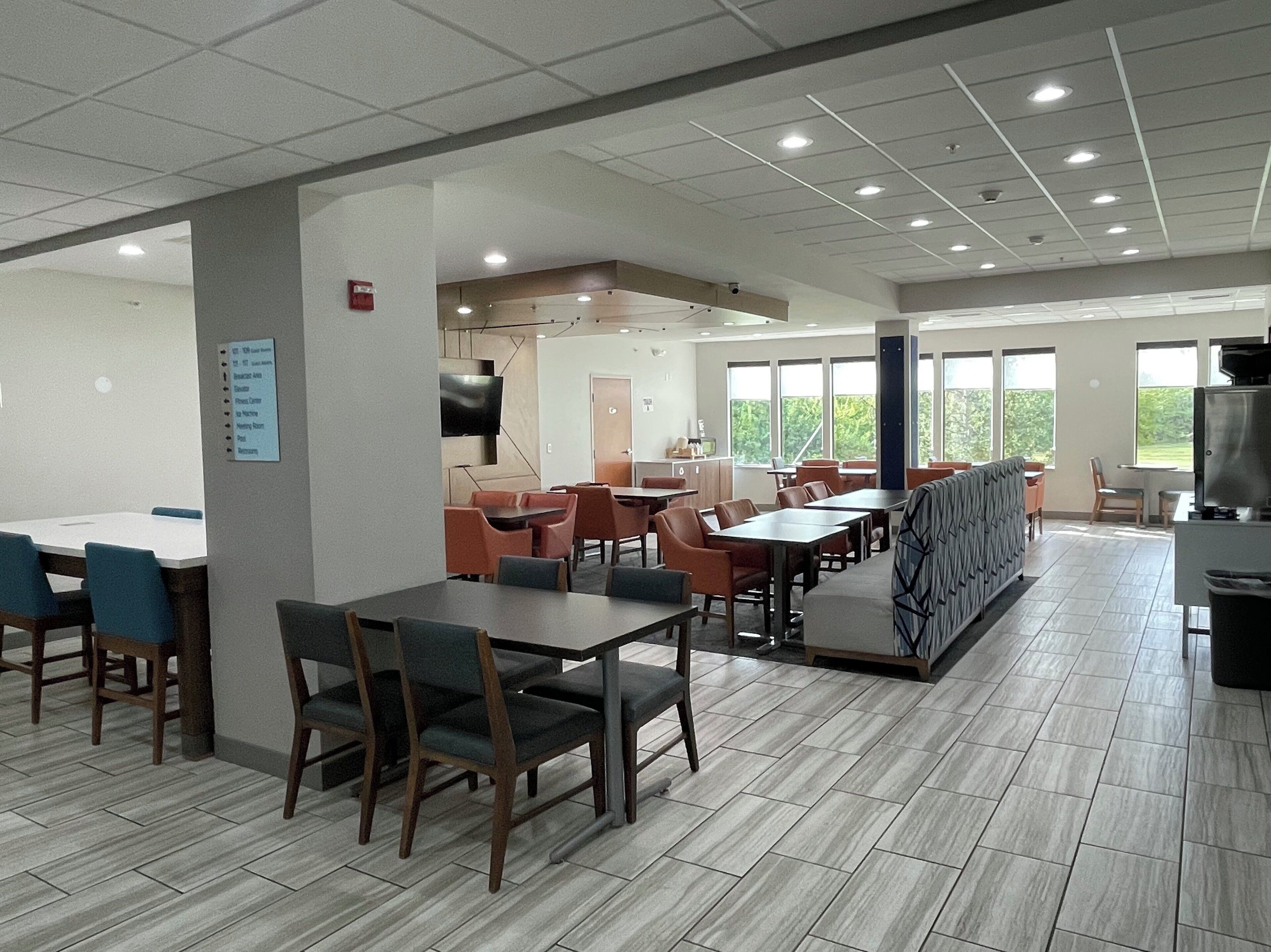 A spacious cafeteria with multiple tables and chairs, a television, and a large window providing natural light.