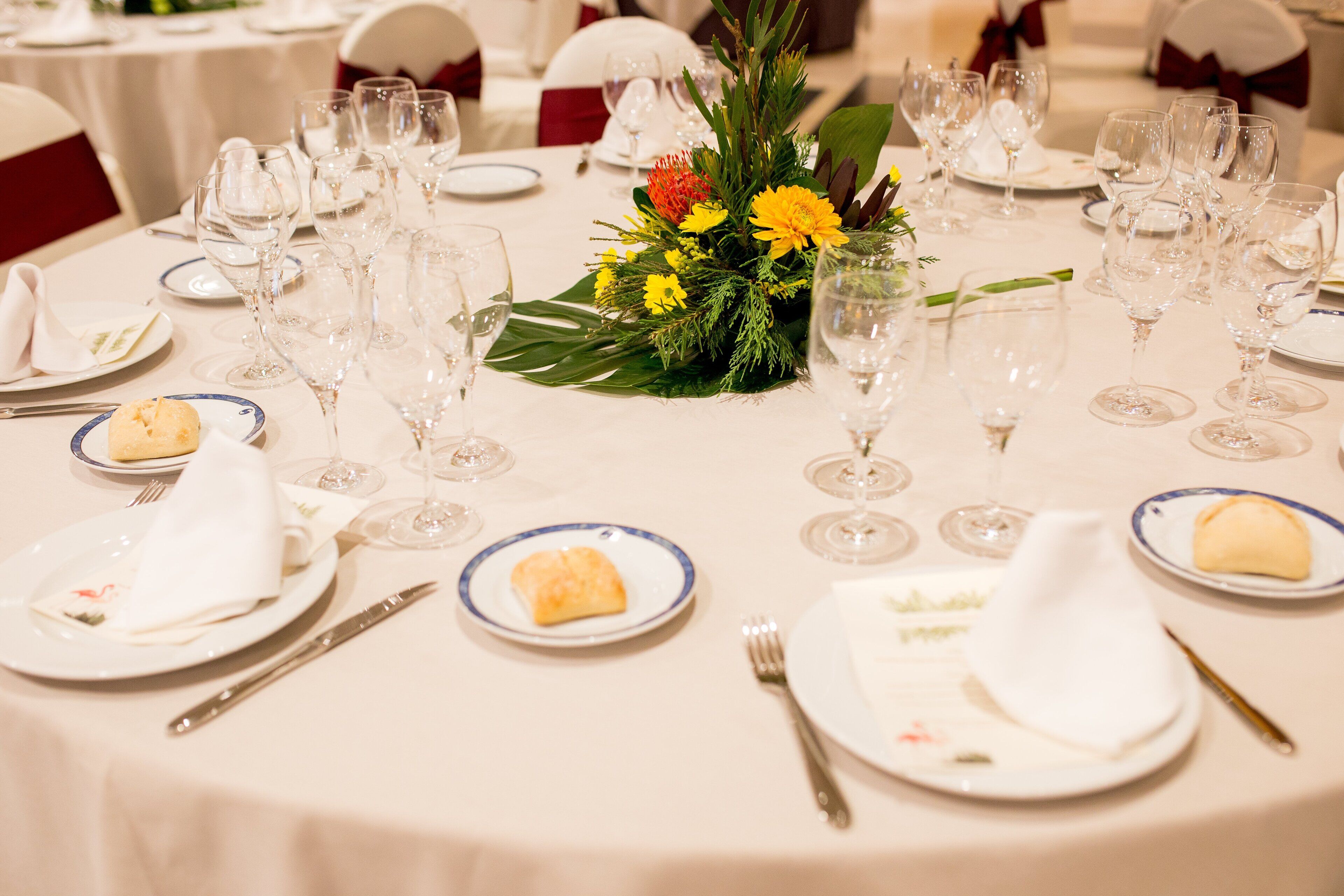 A round dining table set with white plates, clear glasses, and a centerpiece of yellow flowers and greenery.