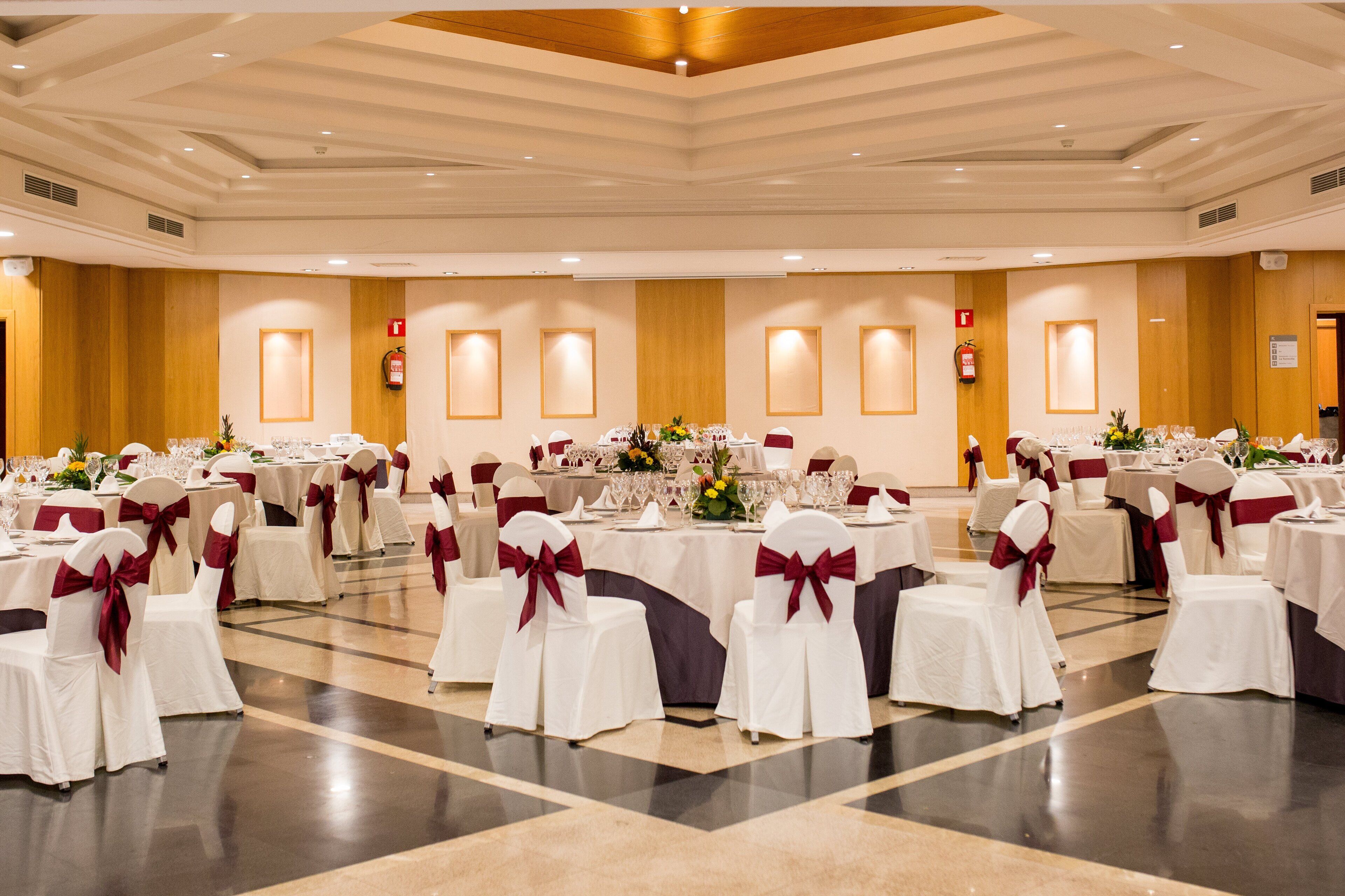 Un salón de banquetes con mesas redondas, sillas blancas con cintas rojas y centros de mesa florales.