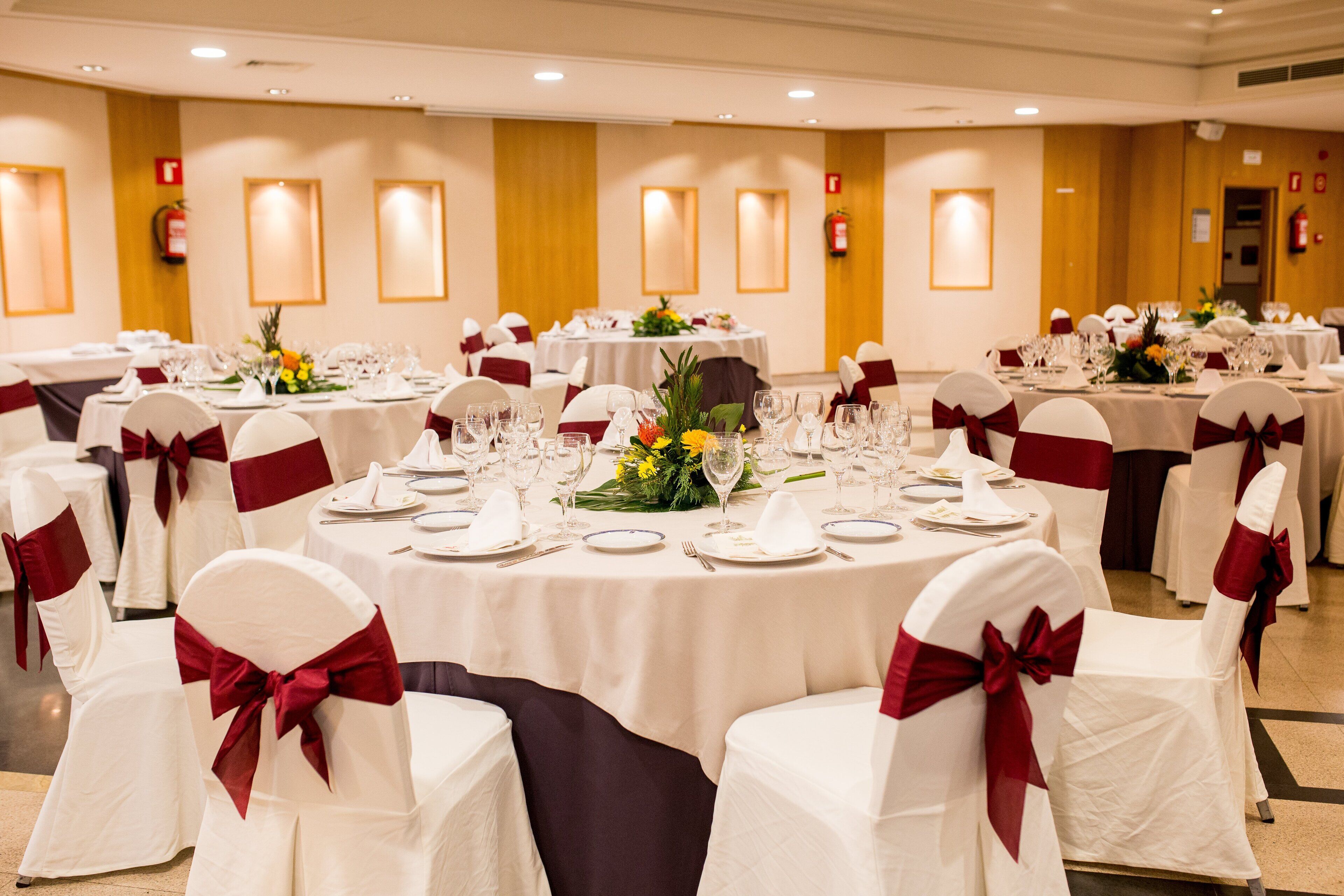 Un salón de banquetes con mesas redondas dispuestas para el servicio de comidas, sillas blancas con cintas rojas y centros de mesa florales.