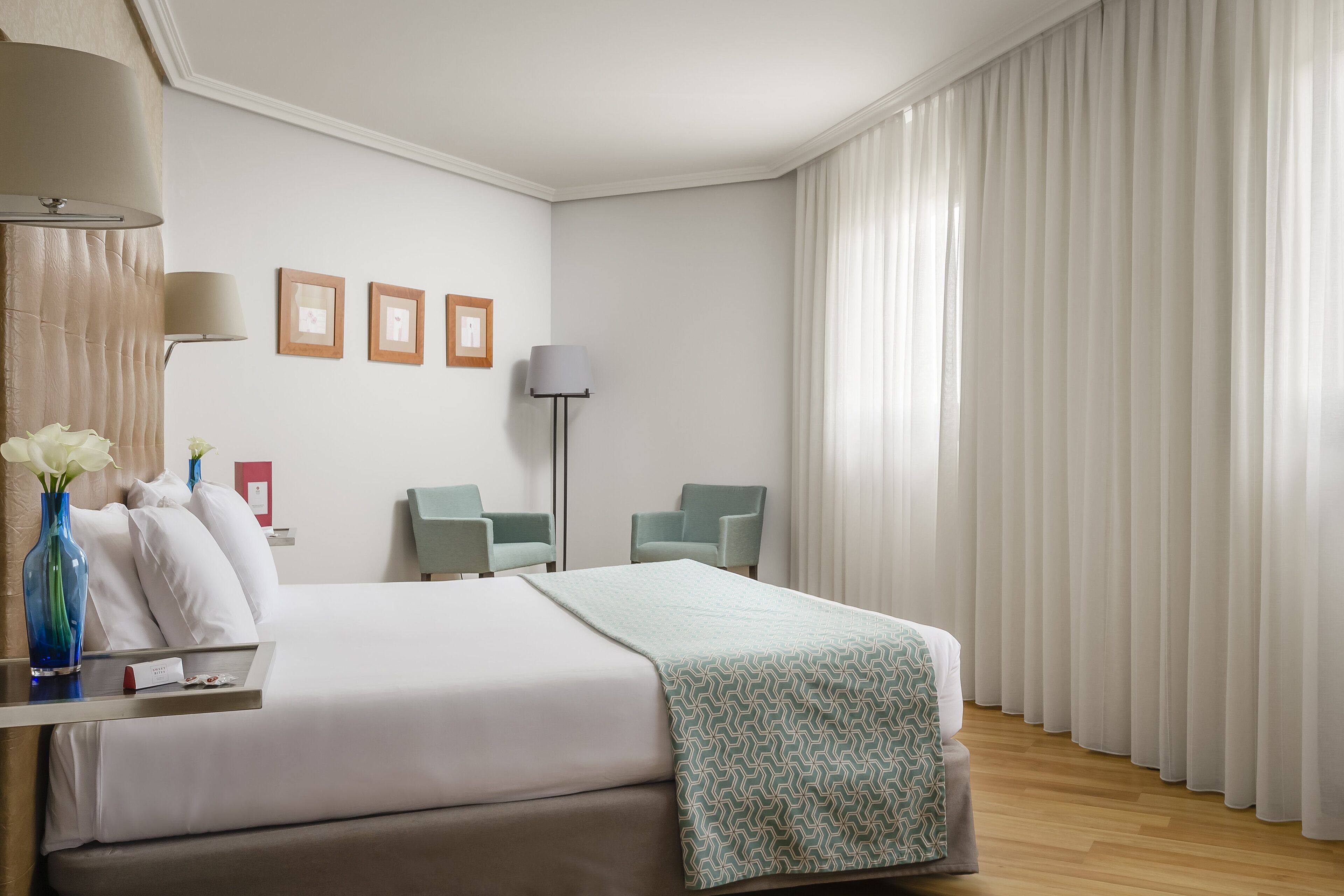 Una habitación de hotel con una cama, dos sillas, un escritorio con lámpara y una ventana con cortinas.