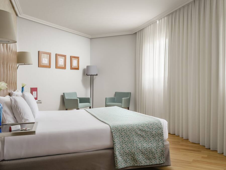Una habitación de hotel con una cama, dos sillas, un escritorio con lámpara y una ventana con cortinas.