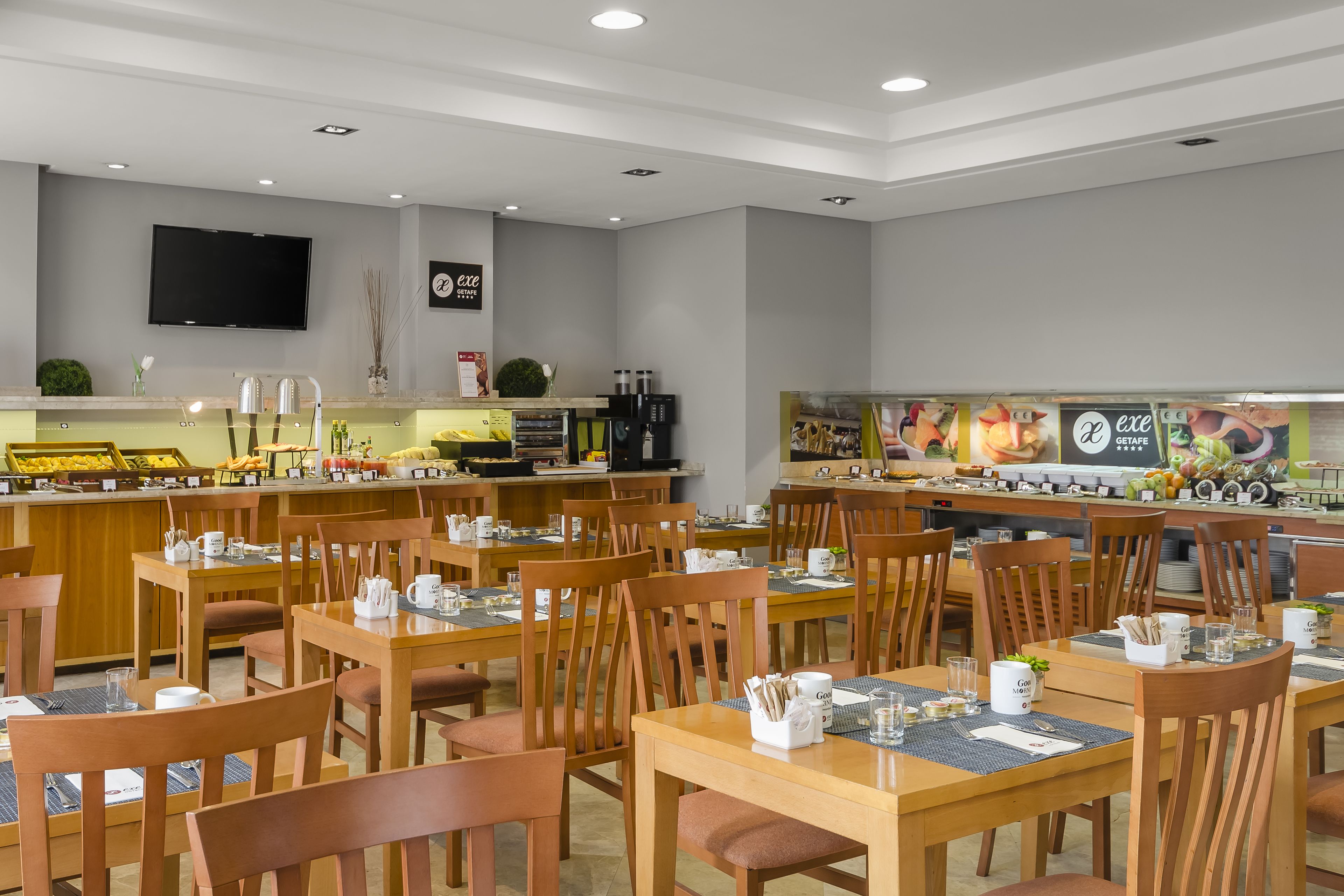 A modern restaurant with wooden tables and chairs, a buffet area, and a flat-screen TV on the wall.
