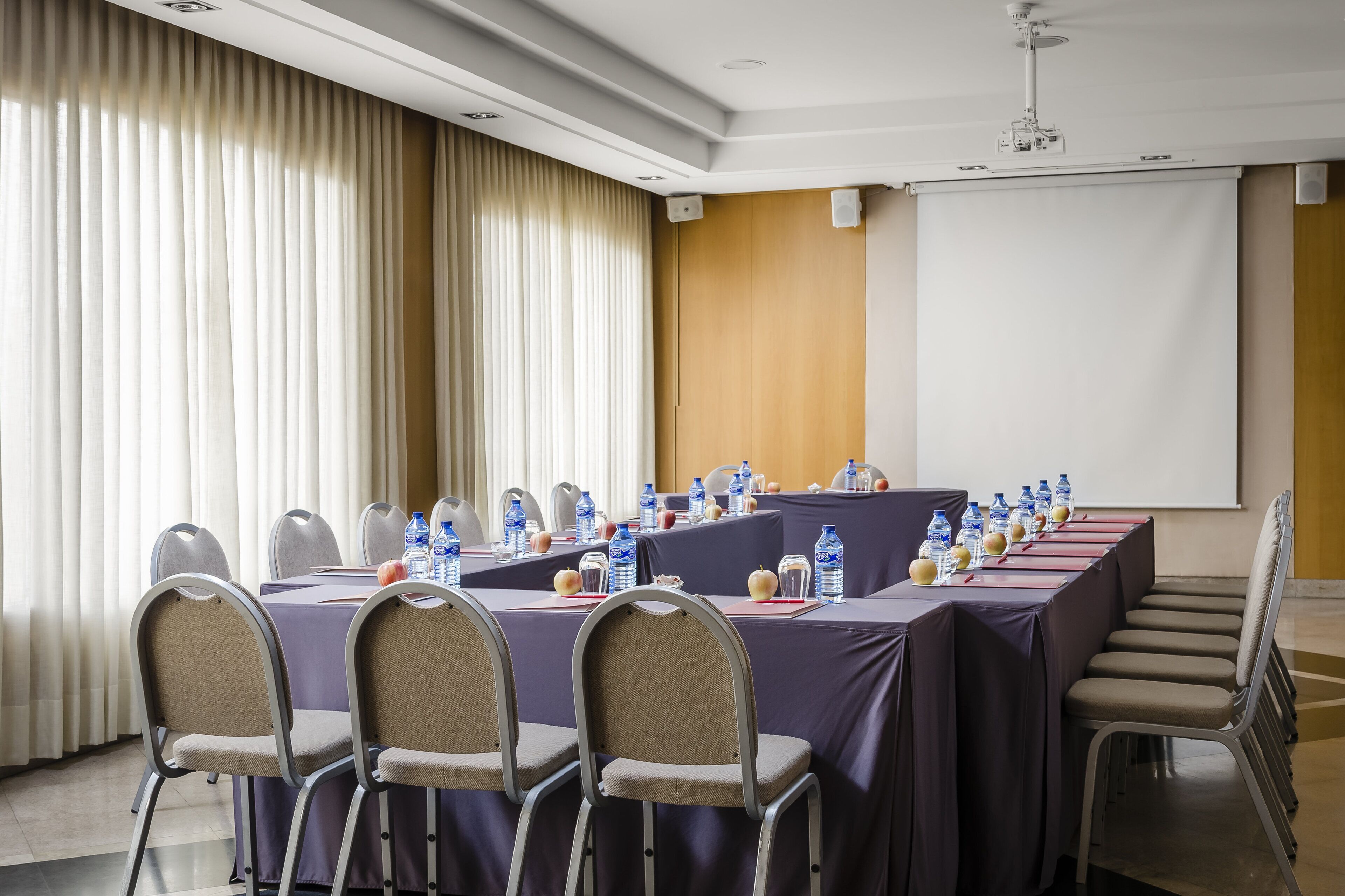 Un salón de conferencias con una larga mesa, sillas, botellas de agua y una pantalla de proyector.