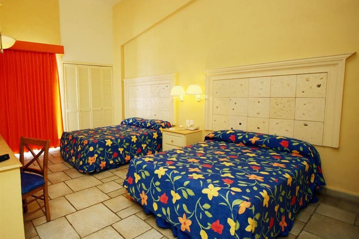 A hotel room with two beds, a desk, a chair, a lamp, and a window with curtains.