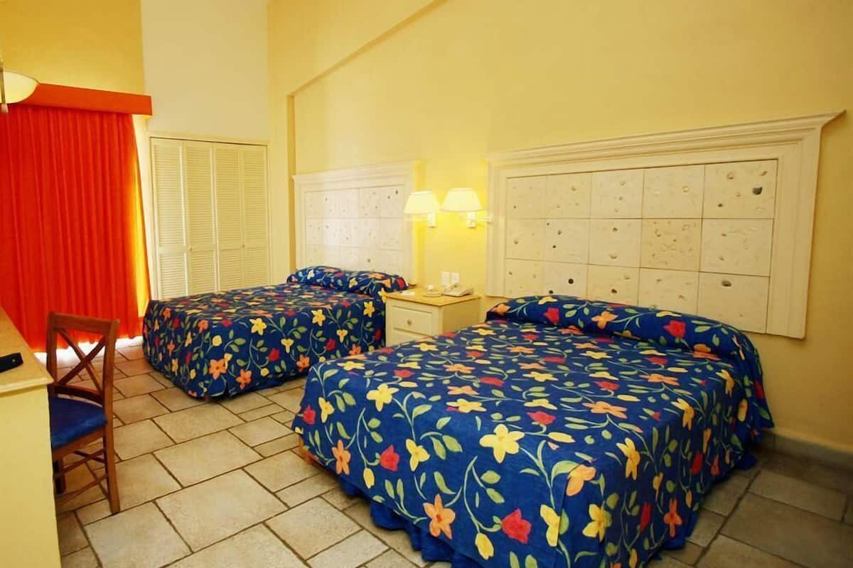 A hotel room with two beds, a desk, a chair, a lamp, and a window with curtains.