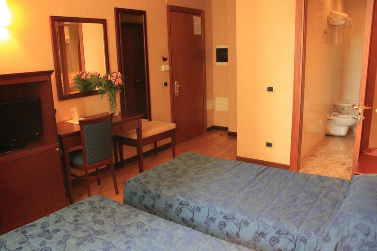 A hotel room with a bed, a desk with a chair, a TV, a mirror, and a bathroom visible through an open door.