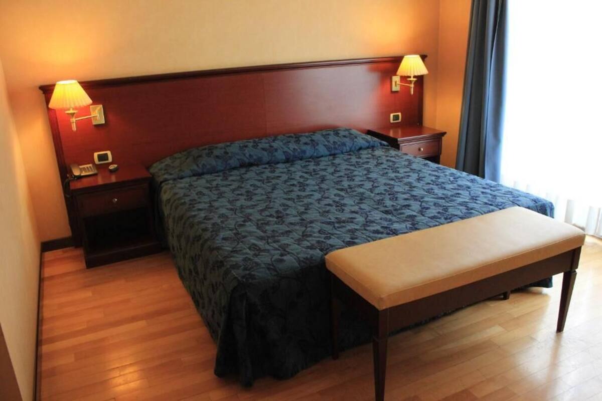 A hotel room with a large bed, a bedside table with a lamp, a bedside phone, and a wooden bench.
