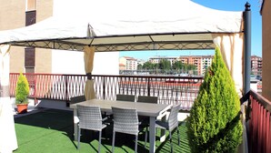 Un patio al aire libre con una mesa y sillas, un toldo blanco y vistas de edificios al fondo.
