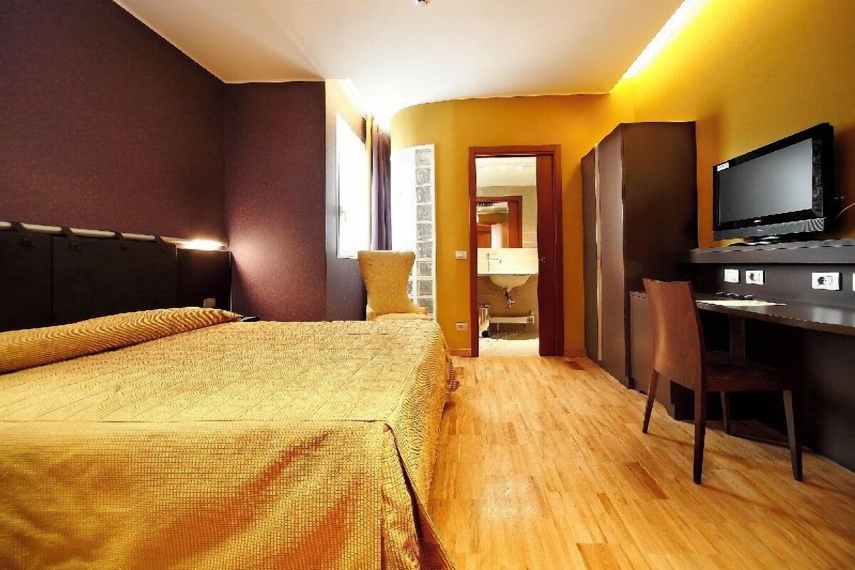 A hotel room with a large bed, a desk with a computer, a television, and a bathroom visible through an open door.