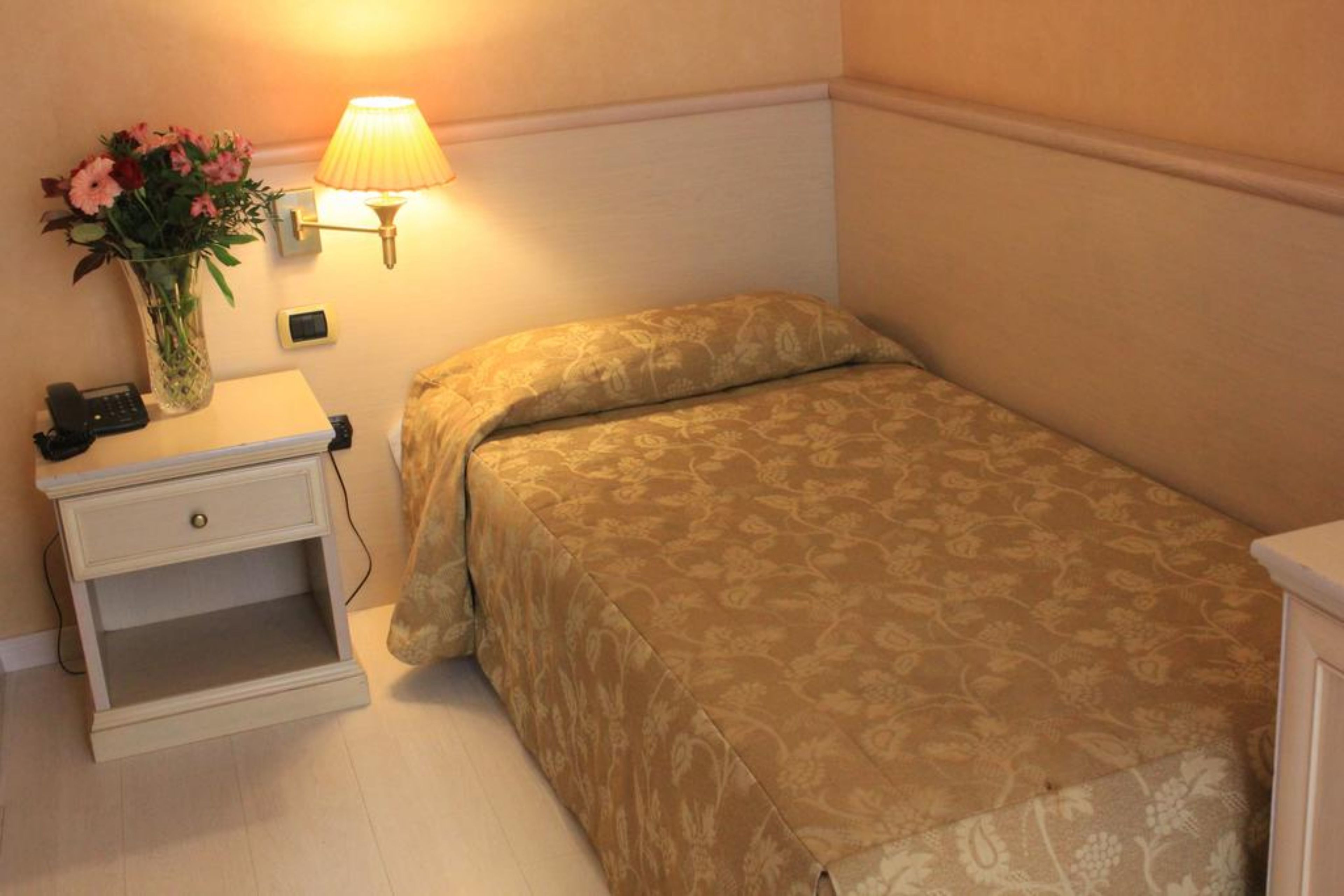 A small bedroom with a single bed, a bedside table with a lamp, and a vase of flowers.