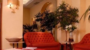 Habitación de hotel con sofá tapizado rojo, colcha roja y una mesa con revistas. Hay una planta en maceta y un jarrón con flores sobre una mesita.
