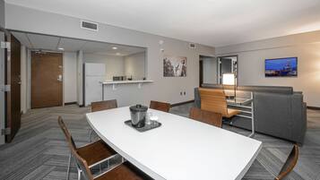 A hotel room with a white dining table, chairs, a kitchenette, a seating area with a sofa and armchairs, and a cityscape view on the wall.