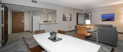 A hotel room with a white dining table, chairs, a kitchenette, a seating area with a sofa and armchairs, and a cityscape view on the wall.