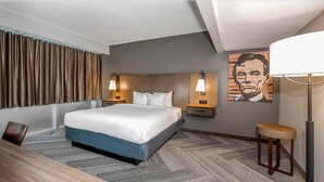 A hotel room with a large bed, two bedside tables with lamps, a wooden desk, a chair, a patterned carpet, and a wall with a portrait.