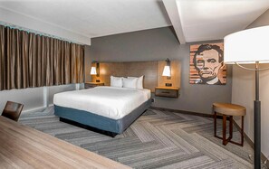A hotel room with a large bed, two bedside tables with lamps, a wooden desk, a chair, a patterned carpet, and a wall with a portrait.