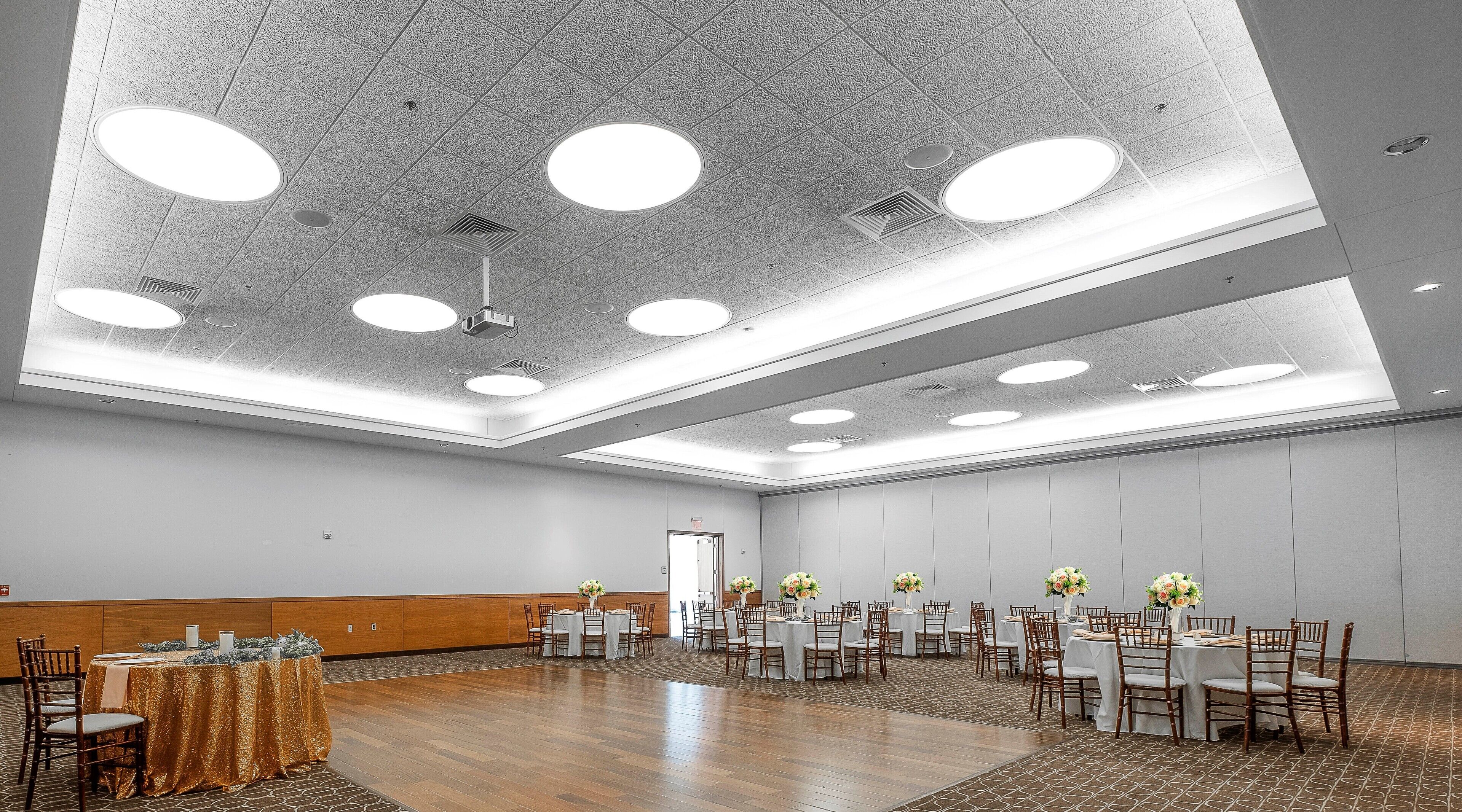Una amplia sala de conferencias con varias mesas y sillas, un panel de madera en la pared y un techo alto con iluminación empotrada.
