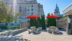 An outdoor seating area with stone tables and red umbrellas, surrounded by trees and a historic building in the background.
