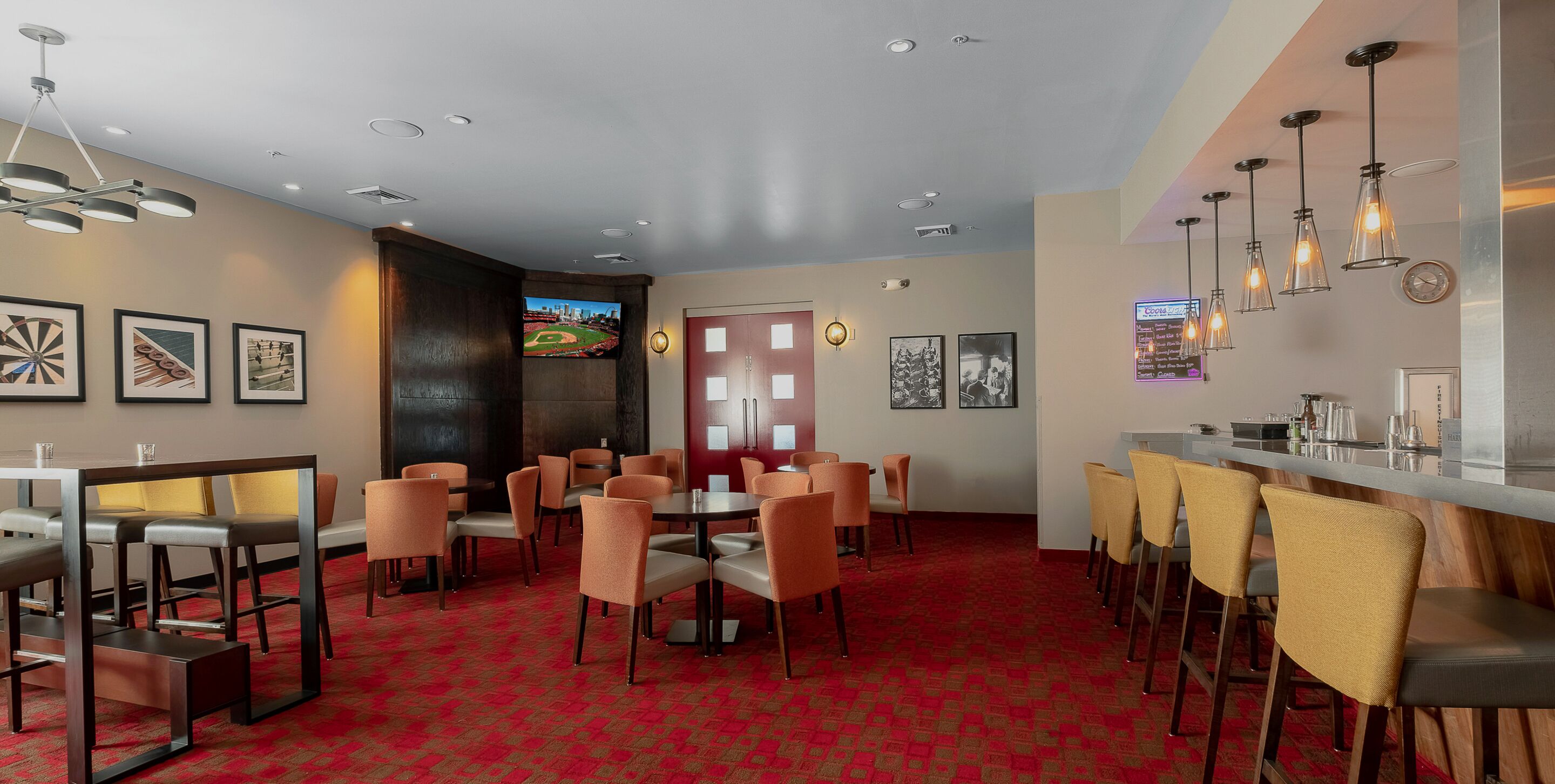A hotel lobby with a red carpet, multiple seating areas, a bar with stools, and a television screen displaying a baseball game.