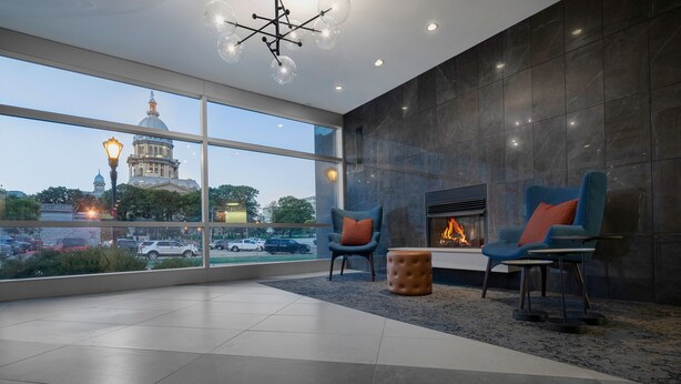 A modern lounge area with a fireplace, two blue armchairs with orange cushions, and a large window offering a view of a cityscape and a dome building.