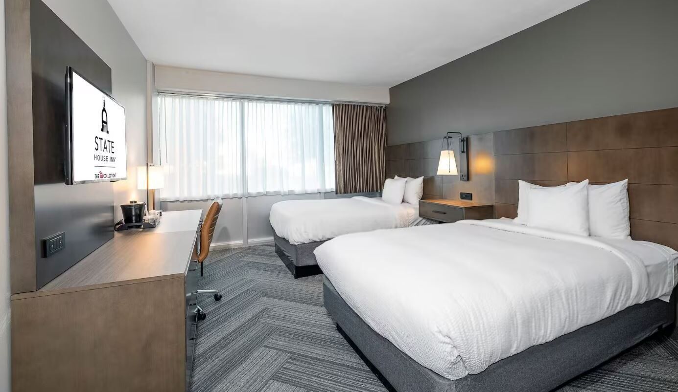 A hotel room with two beds, a desk, a chair, a TV, and a window with curtains.