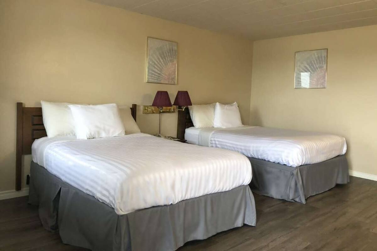 Een hotelkamer met twee eenpersoonsbedden, elk voorzien van een grijze bedrok en witte lakens. Er staan twee wandlampen met paarse lampenkappen.
