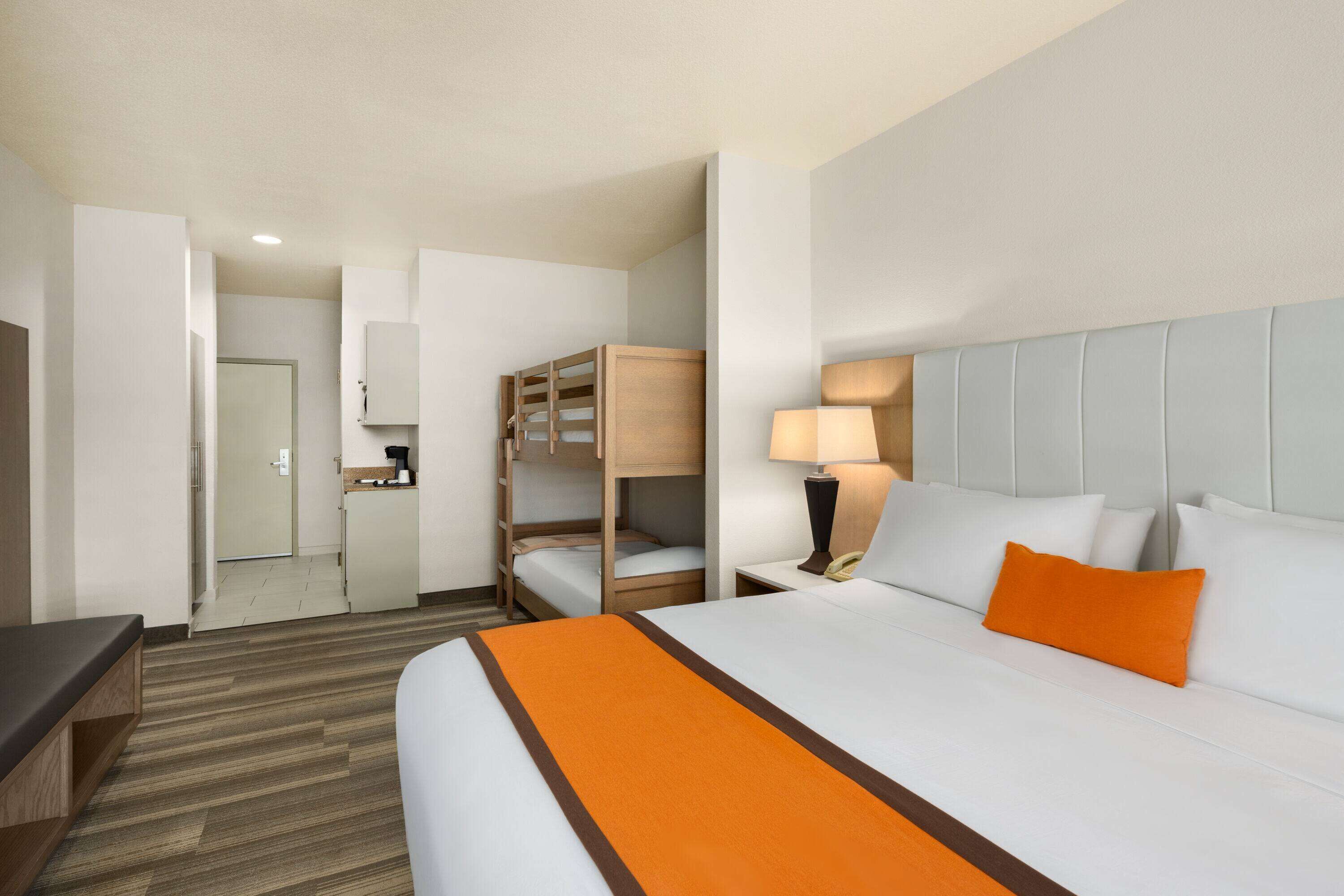 A hotel room with a large bed, a bedside table with a lamp, a bunk bed, and a small kitchenette.