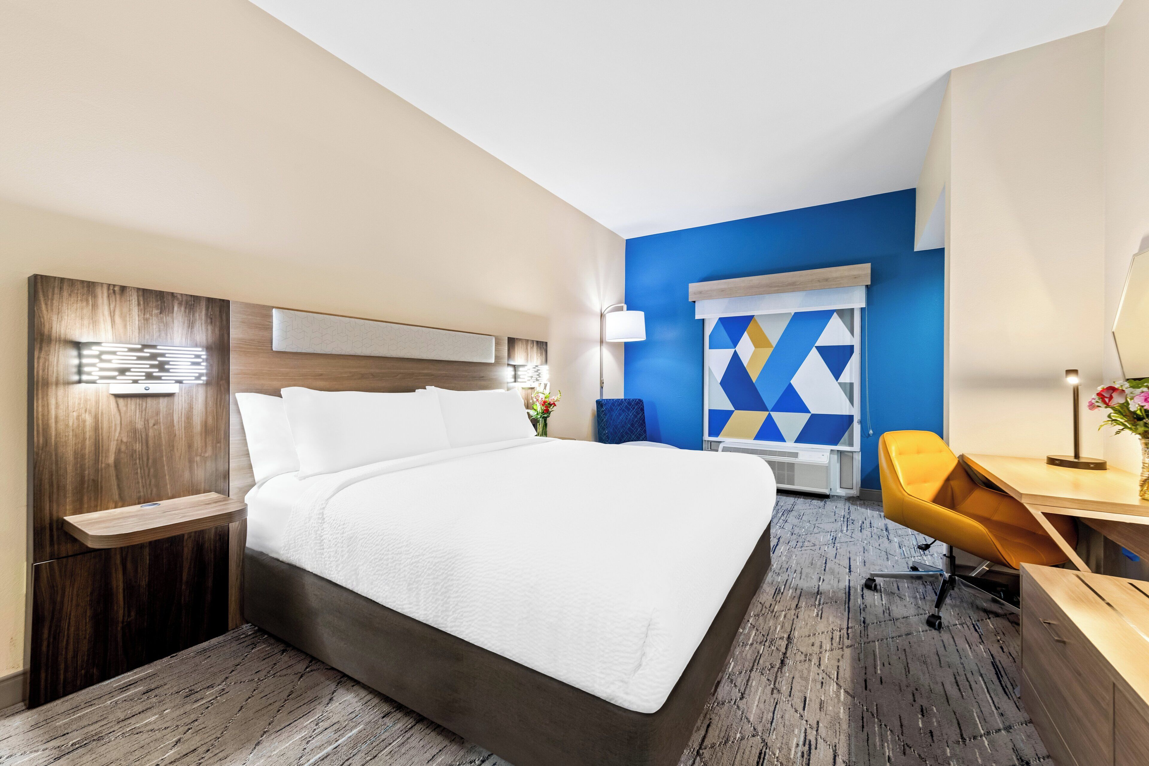 A hotel room with a large bed, a desk, a chair, and a window with a geometric pattern.