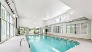 An indoor swimming pool with clear glass walls, a white interior, and a seating area with chairs and a table.