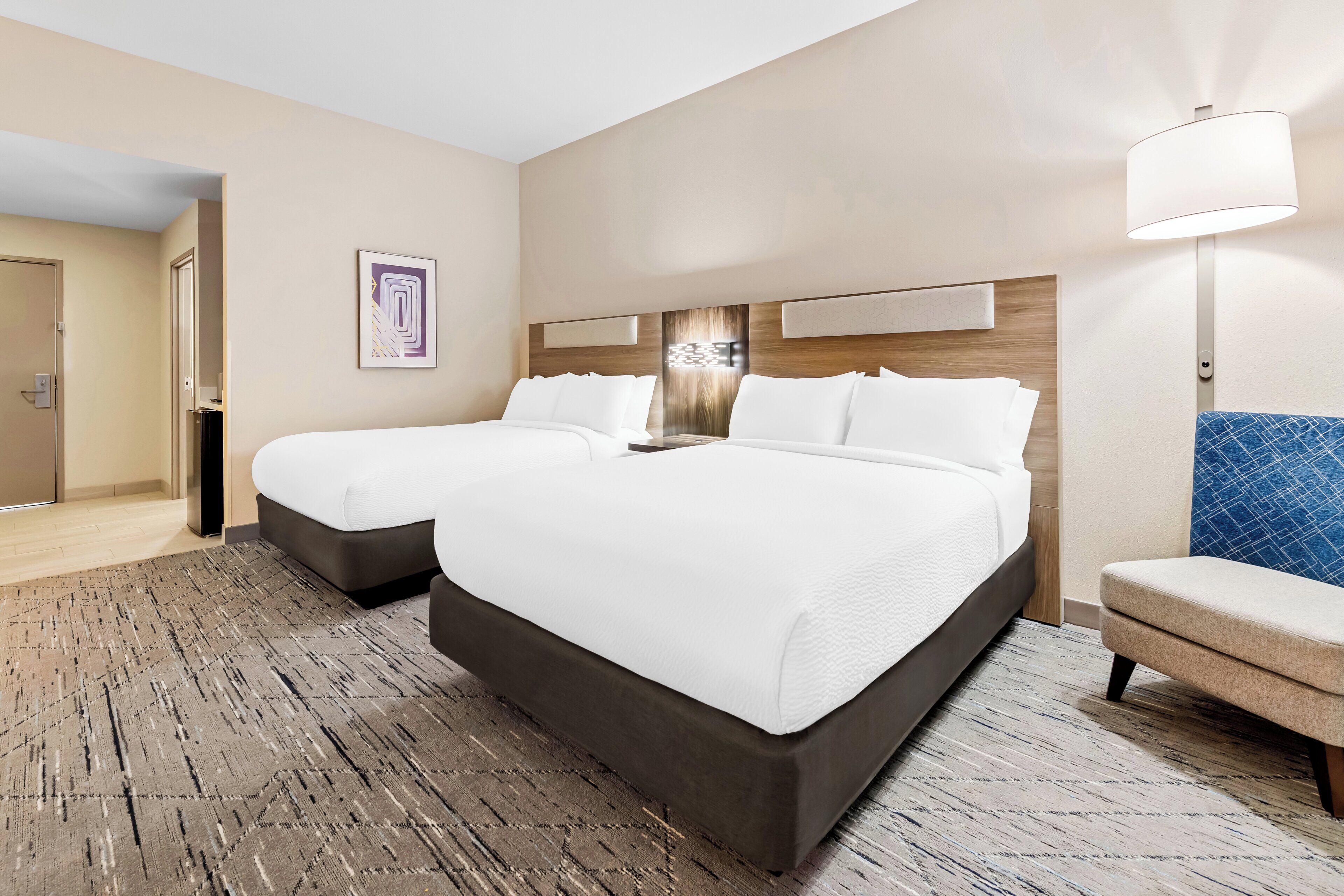 A hotel room with two beds, a desk, a chair, and a lamp.