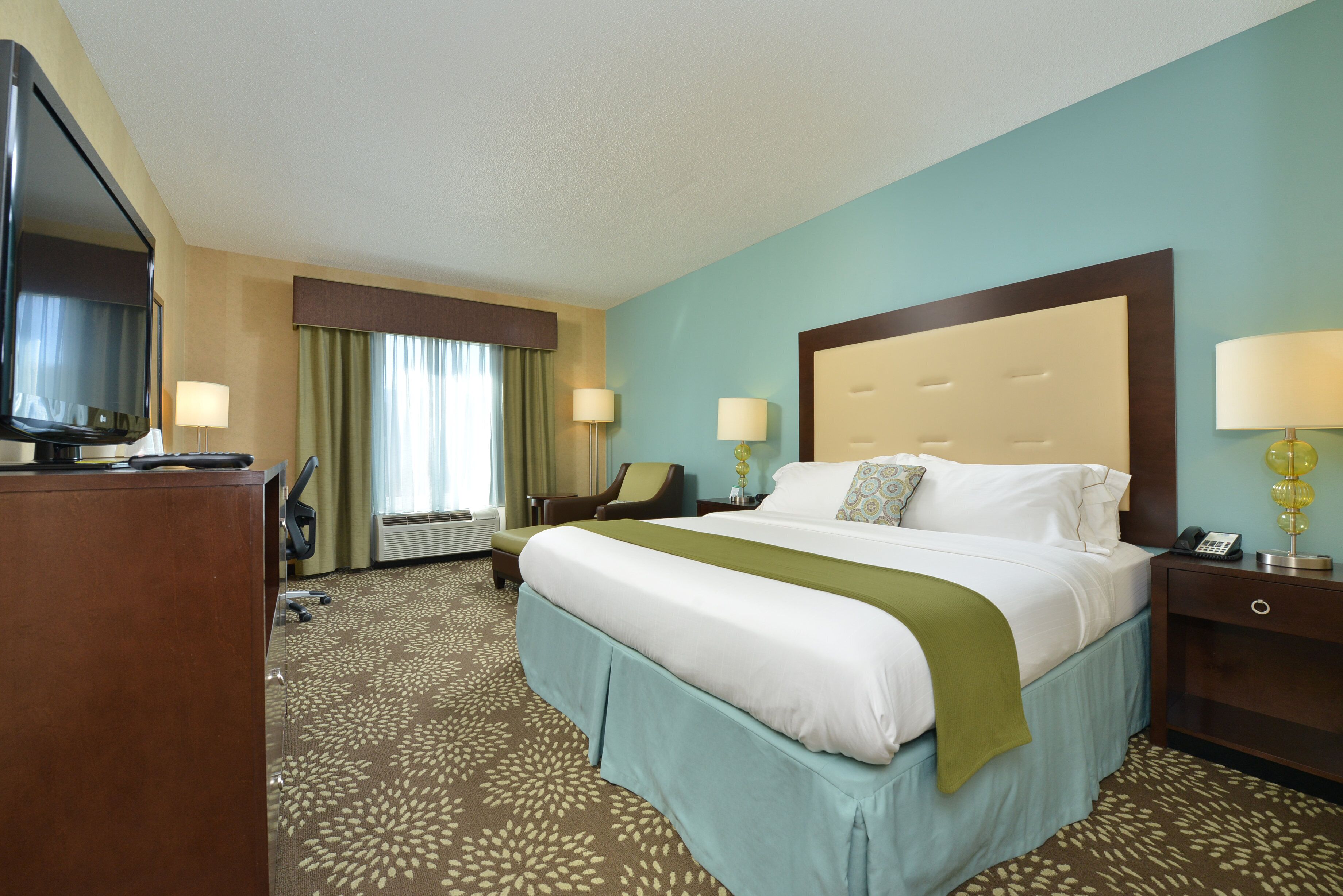 A hotel room with a large bed, two bedside tables, a desk with a chair, a television, and a window with curtains.