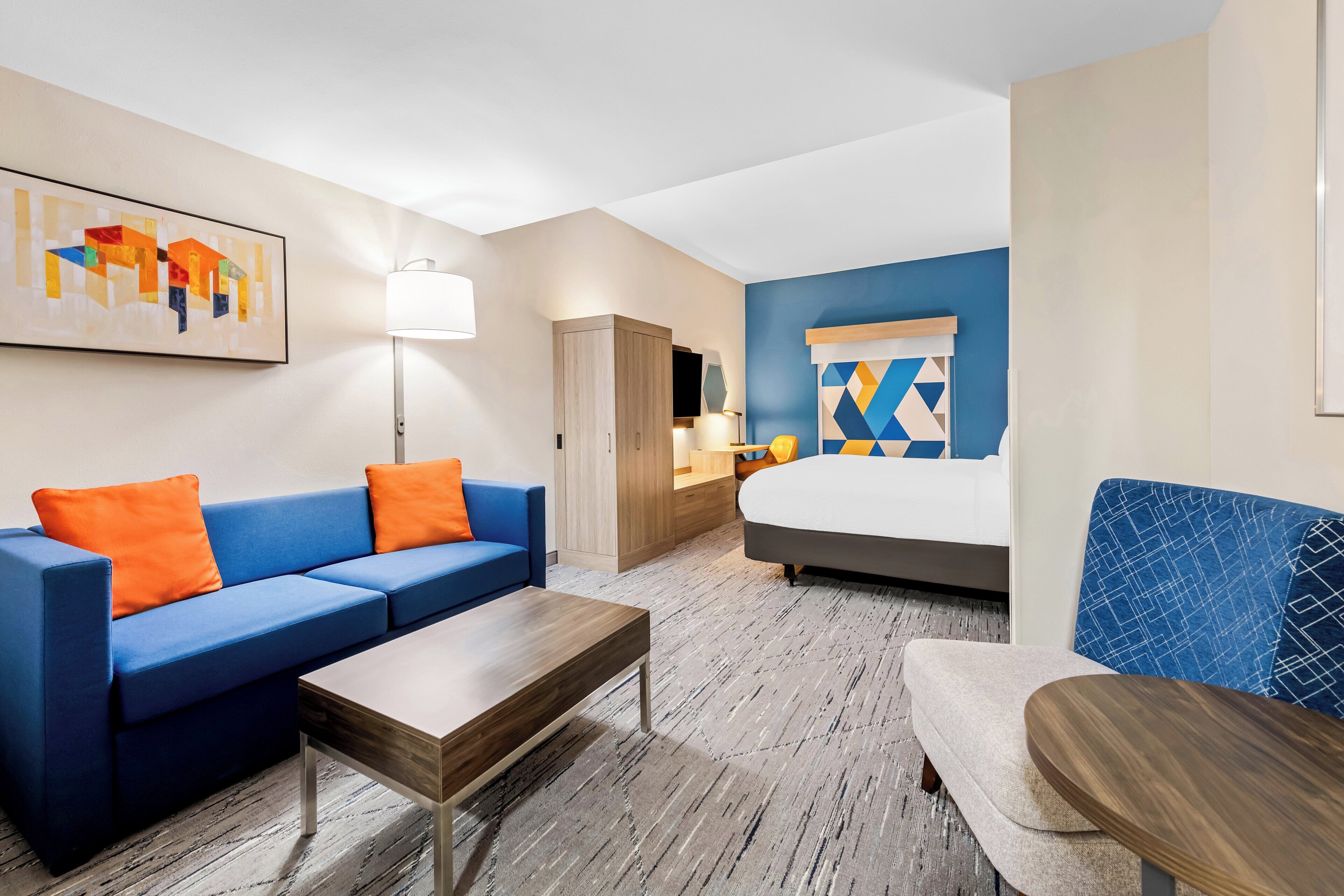 Habitación de hotel con sofá azul, una mesa de centro de madera, cama con cabecera de estampado geométrico, armario y escritorio con lámpara.