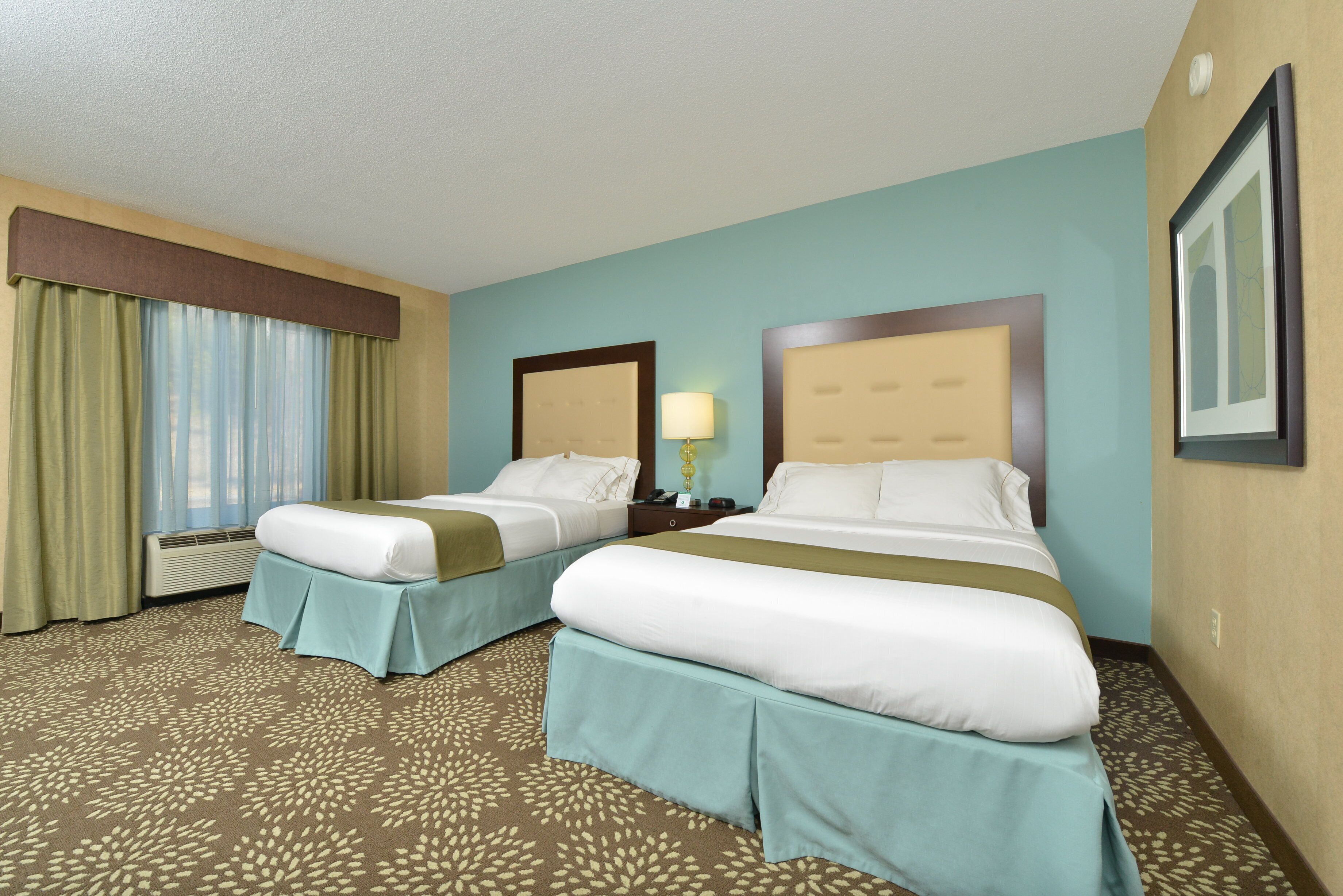 A hotel room with two beds, a window with curtains, a bedside table with a lamp, and a wall-mounted TV.