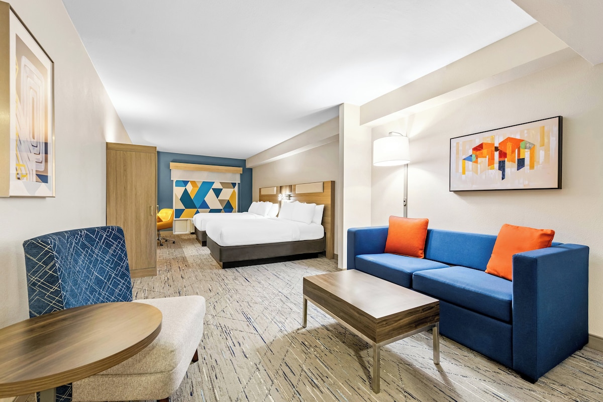 Une chambre d’hôtel avec un lit, un canapé bleu avec des coussins orange, une table basse en bois et une chaise bleue.