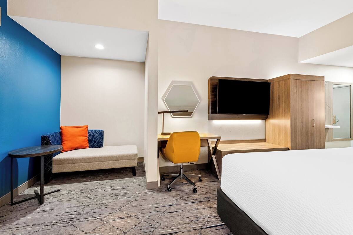 A modern hotel room with a bed, a desk with a chair, a sofa with cushions, a TV mounted on the wall, and a small table.
