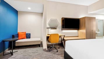 Habitació d'hotel moderna amb llit, escriptori i cadira, sofà amb coixins, televisió penjada a la paret i taula petita.
