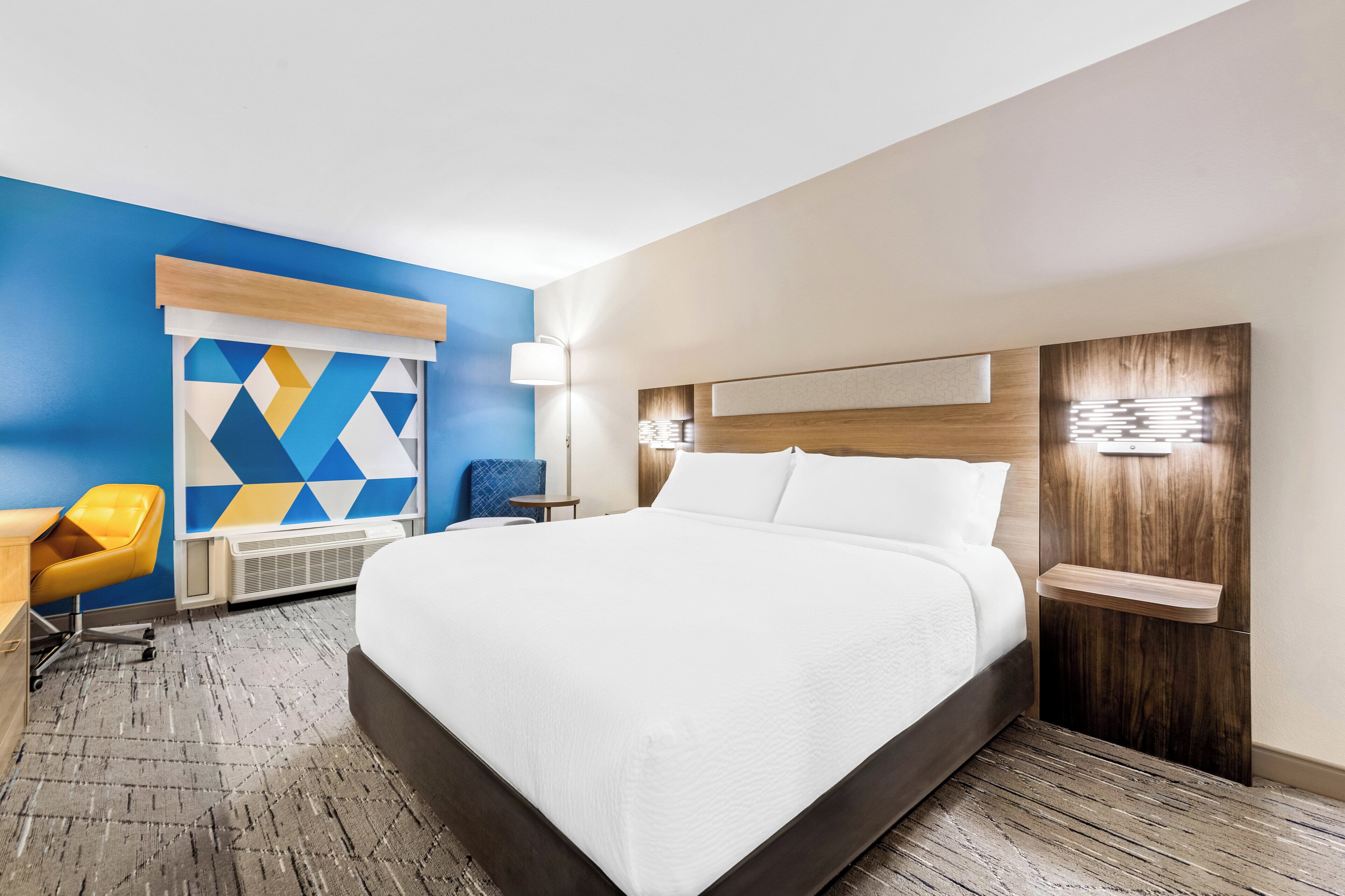 Habitación de hotel con una cama grande, un escritorio con silla, una lámpara de pared y un adorno geométrico en la pared.