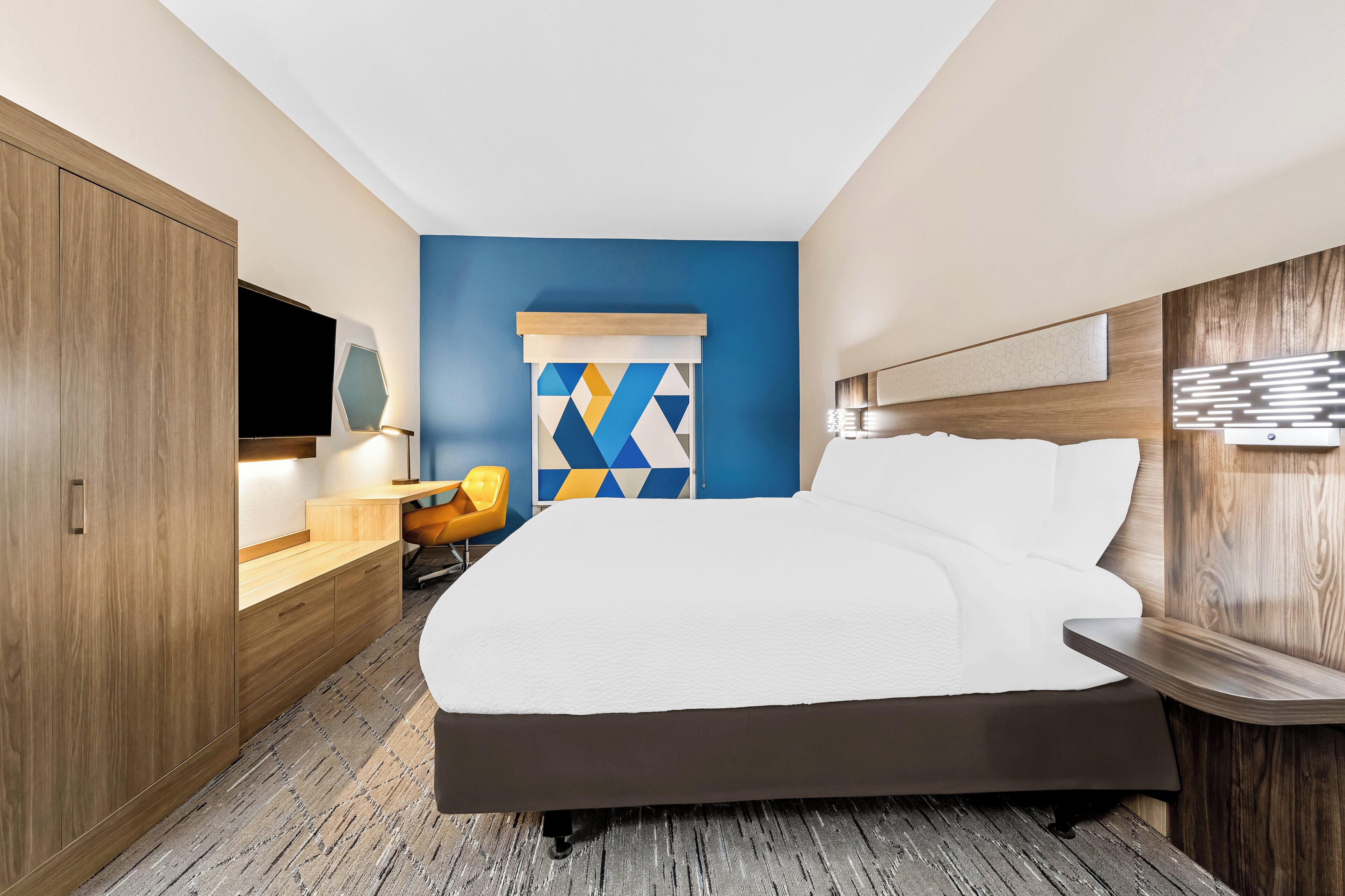 Habitación de hotel moderna con una cama grande, armario de madera, un escritorio con silla y una obra de arte geométrica y colorida en la pared.