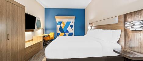 Habitación de hotel moderna con una cama grande, armario de madera, un escritorio con silla y una obra de arte geométrica y colorida en la pared.
