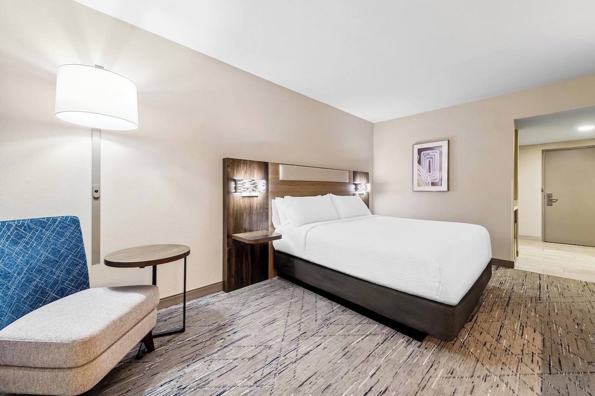 Une chambre d’hôtel avec un lit, une petite table ronde, une chaise bleue, une lampe et un tableau encadré au mur.